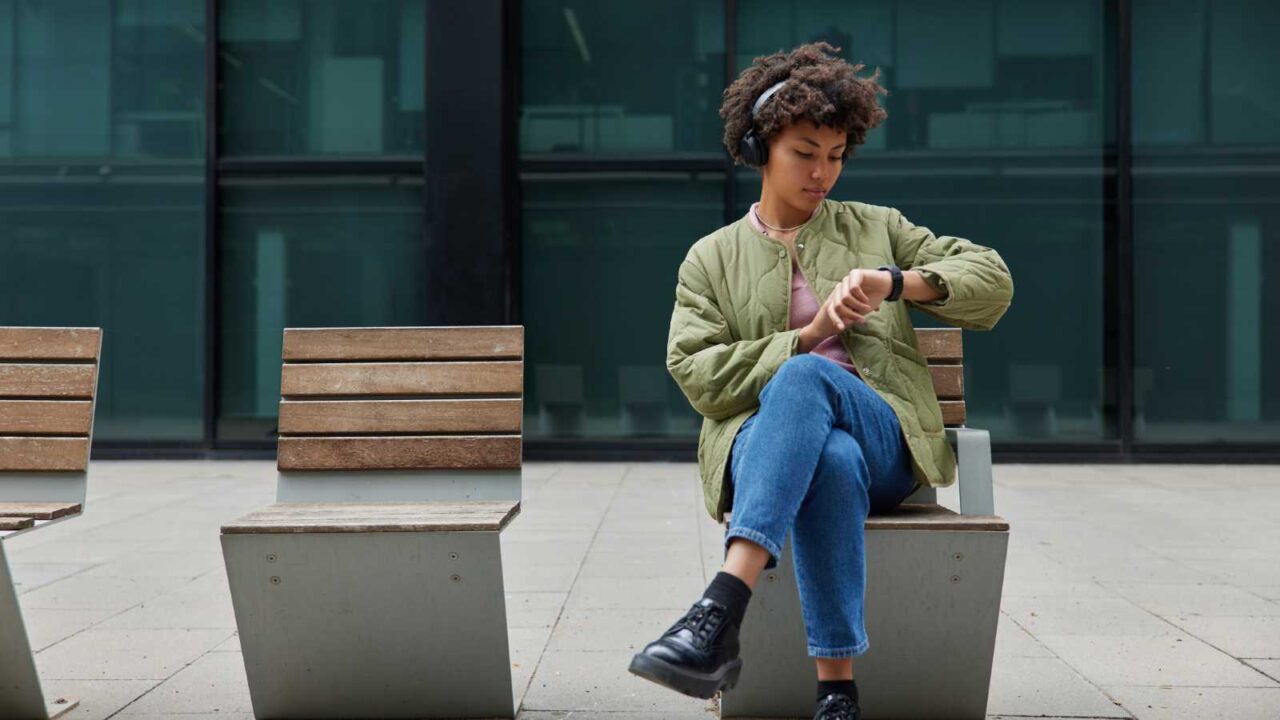 woman wearing headphones while sitting on outside bench waits for someone and looks at smart watch.